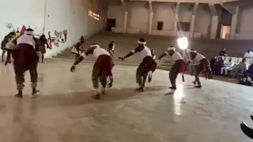 Inside a bright, concrete room, a group of at least eight people perform what appears to be a traditional dance. Seven of the individuals are dressed in similar outfits: white shirts, white headbands, green pants, and maroon fabric tied around their waists. They are performing coordinated movements, leaning forward with their arms outstretched, their bodies slightly bent at the waist. They are facing away from the camera. One additional dancer, dressed in different attire, stands further back on the left side of the frame.

To the right of the dancers, bleacher-style seating rises upward. A small group of people is sitting in the bleachers, watching the performance. A bright light stands in front of the bleachers, illuminating the dancers.

The room has white walls, and a colorful mural is visible on the left side of the frame. The floor is smooth and concrete. The overall setting appears to be an auditorium or community hall.