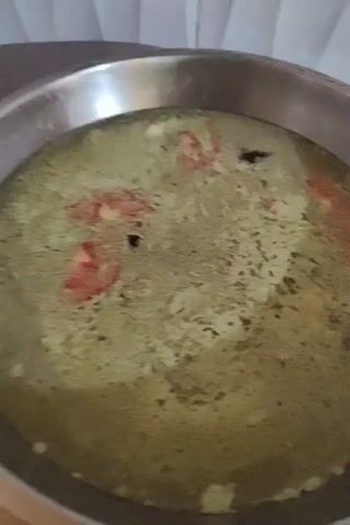Here is a description of the video.

The video shows a wide, shallow, stainless steel cooking pot filled with a murky green broth. Several pieces of red tomato and dark spices float within the liquid. The pot sits on a black stovetop, and the background reveals a white, vertically-ridged surface. The camera briefly pans slightly, capturing the pot's handle and the overall cooking scene.