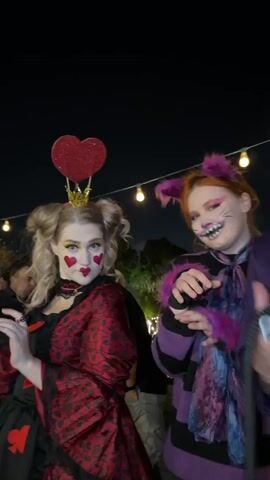 Four individuals in elaborate costumes pose for the camera at what appears to be a night event or party in Nir Eliyahu, Israel.

From left to right:
1.  A person dressed as the Queen of Hearts, wearing a red patterned dress, a crown with red hearts, and red heart makeup on their cheeks.
2.  A person costumed as the Cheshire Cat, with a purple and black striped outfit, cat ears, painted whiskers and nose, and holding a black, whip-like accessory.
3.  A person with blue face paint, green round sunglasses, a purple jacket over a white shirt with a green bow tie and green cummerbund, holding a colorful hookah.
4.  A person dressed as the Mad Hatter, wearing a tall top hat with a clock, dramatic makeup around the eyes, a white ruffled collar, and a purple jacket.

All subjects are smiling and looking towards the camera, conveying a cheerful and playful mood. The background is dark, indicating that the event is taking place at night.