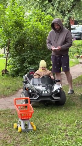 The video shows a fair-skinned man in a brown hoodie and patterned shorts standing on a grassy area, operating a remote control. In front of him, a child wearing a light brown outfit and yellow hat sits in a black toy ATV-like vehicle. Beside the ATV is an orange toy shopping cart. The background includes green bushes, a paved path, and a dark-colored car parked near a brick building. The video then transitions to a selfie perspective of a light-skinned woman with long blonde hair, wearing a green jacket. Behind her, a red toy car is visible on the grass. The camera movement is shaky and erratic throughout the video.