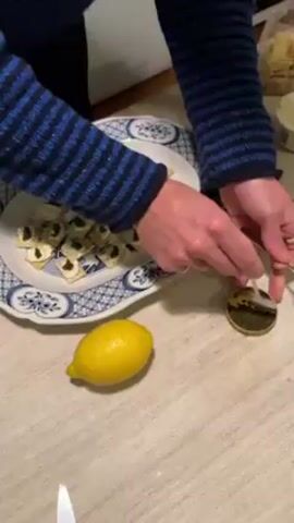 A person in a blue and black sweater spoons caviar from a small gold container onto crackers topped with cream cheese on a white plate with blue floral designs. A lemon sits near the plate. The scene takes place on a light-colored wooden table.