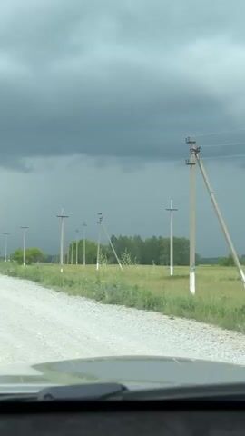 The video is filmed from inside a vehicle, looking out through the windshield at a gravel road stretching into the distance under a dark, overcast sky. The vehicle's dashboard is visible at the bottom of the frame. To the right of the road, a field of green grass and low vegetation extends alongside a row of light-colored utility poles that recede into the distance. Beyond the field and poles, a line of green trees is visible. In the far distance, a heavy curtain of rain or mist descends from the dark clouds, creating a lighter, hazy band across the horizon. A tall, slender white structure, possibly a silo or tower, is faintly visible in the far left distance, partially obscured by the atmospheric conditions. No people or active interactions are visible.