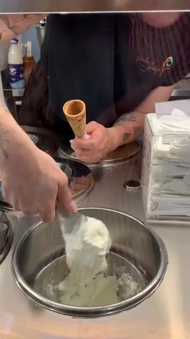 In a close-up shot, a person with visible tattoos on their arms is scooping ice cream into a cone. The person's left hand holds an empty waffle cone, while their right hand uses a silver ice cream scoop to gather a portion of white ice cream from a stainless steel container. The background shows more stainless steel containers with lids, and a dispenser filled with white napkins or utensils. The person is wearing a dark t-shirt with a logo that appears to say "Gelato" and "Felice". The environment seems to be an ice cream shop counter.