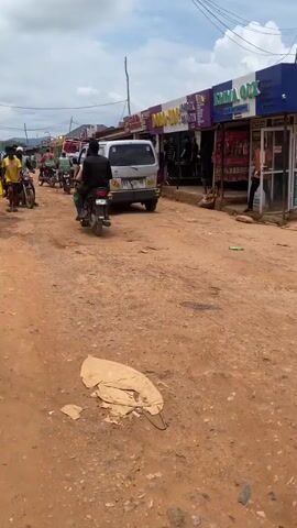 A dirt road is shown with some buildings and people. The road is unpaved and has some potholes. There are several people walking and riding motorcycles. Some of the buildings appear to be shops. There is a white van on the road. The sky is cloudy.