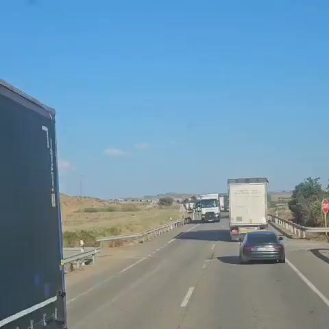 A convoy of trucks and cars travels down a highway on a clear, sunny day. The landscape is dry and dusty, with rolling hills covered in brush and grass. A white semi-truck with "TRANS-ROYP" written on its side is in the foreground, followed by other trucks and cars. In the distance, a line of trucks is parked along the side of the road. A transmission tower is visible on a hill in the background.