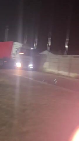 A white semi-truck with red accents is driving at night. The truck's headlights are on, illuminating the road ahead. A concrete fence is visible to the right of the truck, with several tall, thin poles extending upwards behind it. The ground in front of the truck appears to be covered in grass. The scene is dark, suggesting it is nighttime.
