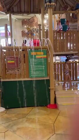 A child in a bathing suit is standing on a wet platform at a water park. Water is spraying from multiple sources onto the floor. In the background, there are water slides and other play structures. The environment appears to be indoors.