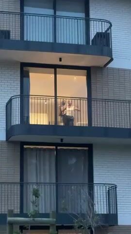 The video shows a woman standing on a balcony of a building. The building is multi-story and made of light-colored brick. The balcony has a black metal railing. The woman is wearing a white tank top and dark pants. She is holding something in her hands and appears to be dancing or moving rhythmically. The interior of the room behind her is lit, showing a minimalist space. There are trees and greenery visible in the foreground.