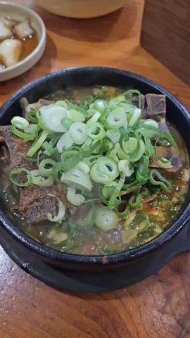 A close-up shot shows a steaming bowl of Korean soup with large pieces of meat and a generous topping of chopped green onions. The soup is served in a dark, round pot. In the blurred background, a small white dish contains sliced white radishes or pickled onions, and a wooden table is visible. The lighting suggests an indoor setting, possibly a restaurant.