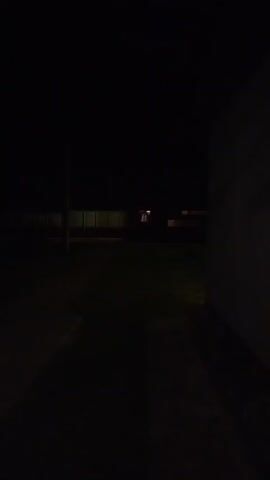 This media file depicts a dark, outdoor scene at night in Krasny Gulyai, Russia. The foreground is dominated by dark shadows, suggesting a path or paved area. In the middle ground, there is a dimly lit grassy area with a faint glow emanating from it, possibly from streetlights or distant building lights. A dark, vertical pole stands on the left side of the frame.

In the background, a long, low structure with several illuminated windows is visible. The overall atmosphere is one of stillness and quiet, with the darkness amplifying the sense of isolation. The time of day is clearly night, and there are no indications of specific weather conditions, though the darkness implies it is not a brightly moonlit night. The location cues are limited to the mention of Krasny Gulyai, Russia.

There are no people or discernible objects besides the structures and the pole. The activity, or lack thereof, is simply a static view of the nighttime environment. The notable details are the stark contrast between the darkness and the faint lights, creating a somewhat eerie or solitary mood. There is no visible text in the media file.