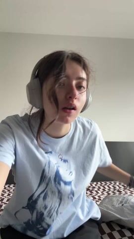 A young woman sitting on a bed wearing headphones is opening a gray plastic package. The package has the word "Öland" printed on it. The woman is wearing a light blue t-shirt with a graphic of a person printed on it. The bed has a brown and white checkered pattern. The wall behind the bed is light gray.