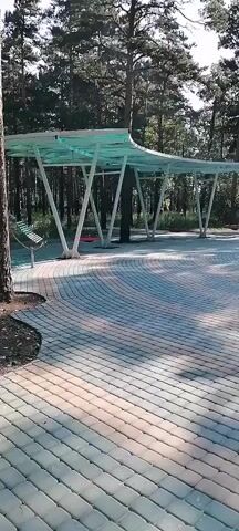 This is a video of a paved outdoor area with trees in the background. There is a covered structure with a translucent roof supported by white pillars. The paving stones are laid in a circular pattern. In the foreground, there is a set of wooden stairs leading up to a stage. The sun casts shadows on the paving stones.