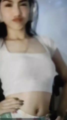 A young woman with long dark hair and headphones is shown from the chest up, wearing a white crop top and patterned shorts. Her left hand is on her hip and she appears to be looking to her left. The background is blurred and appears to be indoors.