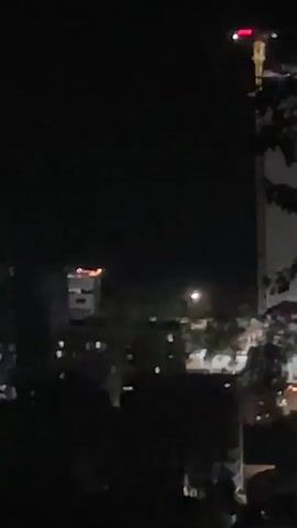 This video shows a nighttime view of a city skyline. It appears to be a time-lapse or a video with rapid transitions between different vantage points, possibly taken from a high-rise building or drone.

The scene features several buildings, some of which are illuminated. There's a prominent skyscraper with a curved, tapered design that is partially lit, with some windows visible. To the left of this skyscraper, there are other buildings with illuminated windows, suggesting they are apartment complexes or offices.

In the center of the frame, there's another building with a distinctive orange-colored light at its top. To the right, further in the distance, another tall, slender building with vertical lighting elements is visible. The lower portion of the video is dominated by dark silhouettes of trees or other structures, indicating the foreground.

The overall impression is one of a bustling urban environment at night, with the lights of the city creating a distinct visual landscape against the dark sky. The lack of discernible human activity or specific events suggests it's a general cityscape view.