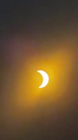 This media file captures a partial solar eclipse. The primary subject is the sun, appearing as a bright, crescent-shaped orb against a darkening sky. The eclipse is in progress, with the moon gradually obscuring a portion of the sun. The lighting is dim, suggesting either early morning, late evening, or overcast conditions, but the focus on the sun's illumination, despite being partially hidden, creates a dramatic visual. The background is a muted gradient of brown and grey, which transitions to black as the eclipse progresses further into the clip.

The activity taking place is a celestial event, a solar eclipse, which is a natural phenomenon. There are no people or man-made objects visible in the frame. The setting appears to be outdoors, given the direct view of the sun. The notable detail is the progression of the eclipse, showing the crescent shape of the sun becoming thinner over time. The overall mood conveyed is one of awe and observation of a natural wonder. There is no visible text in the media file. The location context of Waters Upton, United Kingdom, implies this observation took place in that region.