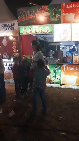 The video captures a nighttime scene at an outdoor market or fair. In the foreground, several food stalls are visible, with signs for "Milo Malaysia" and "Thai Tea." People are gathered around the stalls, with some appearing to be ordering or waiting for food. One stall has a large banner advertising "Cheese Coin." Further back, a Ferris wheel or similar amusement ride is illuminated with colorful lights and is in motion, with people riding it. The ground appears to be dirt or sand, with scattered debris like trash. The overall atmosphere is one of a lively public gathering.