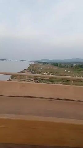 The video shows a view from a moving vehicle on a bridge. The bridge has a tan-colored concrete barrier and a gray metal railing. To the left, the view is dominated by a wide river, stretching to the horizon where another, longer bridge is visible. On the right bank of the river, the land is mostly dry and grassy, with some sparse vegetation and a few trees. In the distance, there are low hills. The sky is overcast, creating a gray and muted atmosphere.