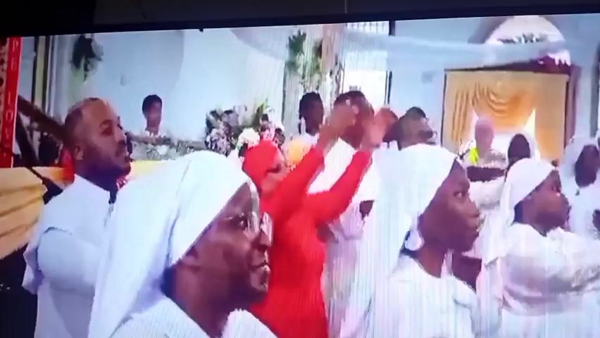 The video displays an indoor event, possibly a religious service or a gathering. Several people are visible, many wearing white attire and head coverings, suggesting a uniform or specific dress code. In the foreground, to the left, a man with glasses is looking forward. To his right, a person in a white veil and glasses is facing away from the camera. To the right of that person, another individual in a white veil is partially visible.

The background reveals a stage or raised platform area. A man in a red, long-sleeved shirt is standing at a podium, speaking into a microphone and looking down at papers. Behind him, a banner with red and white patterns and text that appears to read "BCS HOPE LOVE" is displayed. Floral arrangements are present on the platform, with white and yellow flowers. More people in white are visible in the background, some appearing to be seated and others standing. The environment seems to be decorated with white fabric draped from the ceiling. The lighting is somewhat dim, contributing to a soft, ambient atmosphere. The overall impression is one of a formal gathering with a speaker addressing the attendees.