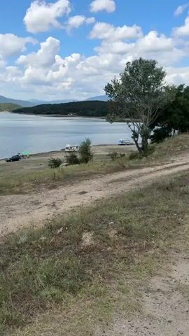 The video pans across a body of water, likely a lake, with a treeline in the background. In the foreground, a dirt path leads down to a sandy shore where two white and blue campers are parked. A lone stork stands on the shore, near a small tree. The scene is outdoors, under a cloudy sky.