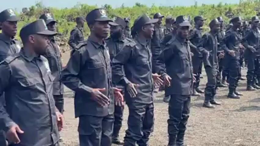 Here is a forensic description of the video:

A group of uniformed men are standing in formation, clapping and singing. They are dressed in dark uniforms with matching caps. The caps have a badge on the front. The men appear to be outdoors, in a grassy area with trees visible in the background. The sky is overcast, and the lighting is somewhat flat. The men are facing towards the camera and clapping in sync. The man in the front row on the right side of the frame is singing.