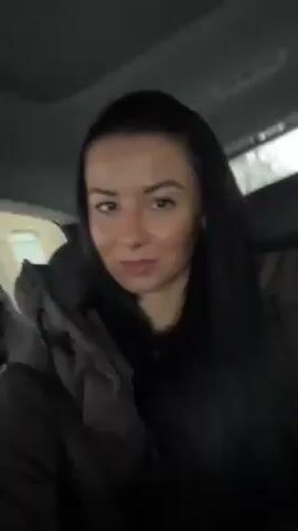A woman with long, dark hair and a tan complexion is seated in the back of a car. She is wearing a dark brown puffer jacket. The car's interior is visible, including the seats and the sunroof. The camera angle is slightly low, looking up at the woman. The lighting suggests it's daytime, but possibly overcast. There is no significant action or interaction in the frame.