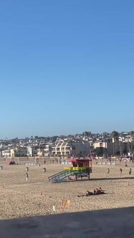 The video shows a beach scene on a clear day. A lifeguard tower painted in rainbow colors stands on the sand. Several people are scattered across the beach, some sitting and relaxing, others standing or walking. In the background, there is a line of buildings, palm trees, and tents set up on the sand, suggesting a possible event or market. The sky is a clear blue.