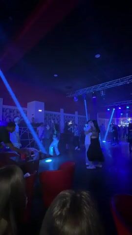 The scene takes place inside a dimly lit nightclub, characterized by blue and red lighting, presumably from stage lights. There's a DJ stand in the background, and a small dance floor where people are dancing in pairs. Several red chairs are visible in the foreground, appearing to be part of a lounge area. The overall atmosphere suggests a casual evening of socializing and dancing.