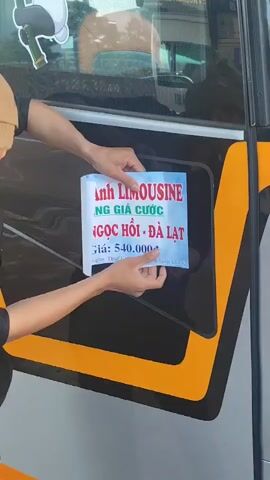 A person wearing a tan baseball cap and a black shirt is affixing a white and blue sign to the tinted window of a bus. The sign, which has red and blue lettering, appears to be an advertisement. The bus has a black and yellow stripe along its side. The background suggests an outdoor setting, possibly a street or parking area. The person's hands are visible as they carefully place the sign on the glass.