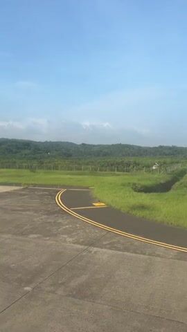 The video pans right across an airport tarmac. In the foreground is concrete with yellow markings. Beyond the tarmac, there is a strip of green grass with a fence running along it. In the background, a dense, tree-covered hill under a bright blue sky with scattered white clouds.