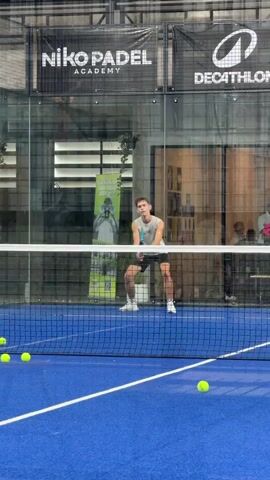 A young man in athletic wear is playing padel on an indoor court. He is in the process of hitting a yellow ball with a padel racket. Several other yellow balls are scattered on the blue court surface. The court is enclosed by glass walls, and in the background, there is signage for "Niko Padel Academy" and "Decathlon". The lighting appears to be artificial, typical of an indoor sports facility. The man is focused on the game, with his body positioned for the shot.