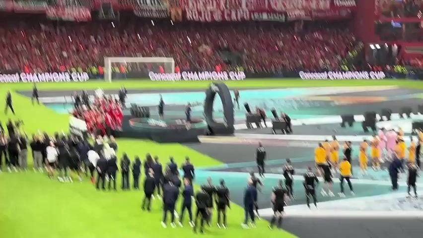 A soccer stadium is packed with people and fireworks are going off on the field. The stands are predominantly red, with banners hung along the top edge. On the field, a large stage has been set up with an archway and the words "CONCACAF CHAMPIONS CUP" displayed. Two groups of people stand on either side of the stage: one group is wearing red uniforms and the other is wearing yellow and black uniforms. Several individuals in suits are also present on the field, overseeing the event. Fireworks are erupting from behind the stage, sending showers of sparks and smoke into the air. The overall scene suggests a celebration or award ceremony following a soccer game.