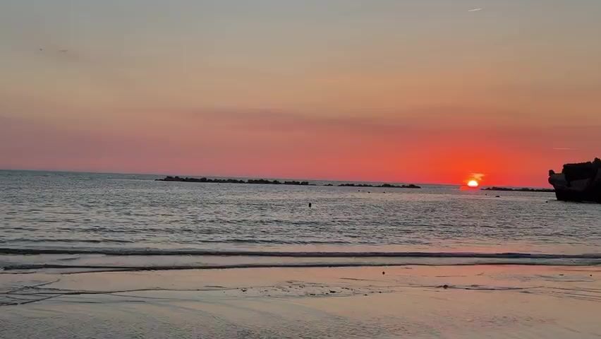 The video shows a wide shot of a calm ocean at sunset. The sun, a bright orange disk, is low on the horizon, casting a reddish glow across the sky and water. The waves are small, gently lapping at the sandy shore in the foreground. In the distance, several rows of dark, rocky structures extend from the water, likely breakwaters. To the right, a dark, silhouetted object, possibly a shipwreck or large rock formation, juts out from the water. A few small birds are visible in the sky and water. The overall atmosphere is serene and tranquil.