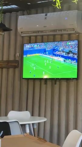A security camera captures a view of a television screen displaying a soccer match. The television is mounted on a wall with vertical wooden paneling. An air conditioning unit is above the television. In the foreground, parts of tables and chairs are visible, suggesting the setting is an indoor space like a cafe or restaurant. The soccer game is in progress, with players on the green field and a net visible on the screen. The lighting is dim, typical of an indoor environment with artificial lighting.