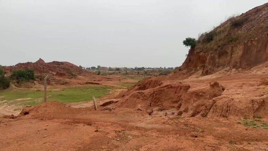 The scene is an outdoor landscape featuring a lot of red soil. There are mounds and ridges of red earth, with some patches of green grass in between. In the background, there are trees and some buildings visible on the horizon, suggesting a nearby town or settlement. The sky is overcast and grey. There are a few wooden or metal posts standing in the scene, seemingly marking boundaries. There doesn't appear to be any human activity in the frame.