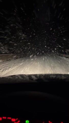 The video, shot from the perspective of a driver, depicts a car navigating a snowy road at night. The foreground is dominated by the windshield, which is partially obscured by accumulated snow or ice at its base, hinting at the harsh winter conditions. Beyond the windshield, the scene is dark, with the car's headlights cutting through the darkness, illuminating the road directly in front.

Heavy snowfall is clearly visible, with countless snowflakes appearing as bright, white specks and streaks against the black background, illuminated by the headlights. They seem to be falling or blowing towards the car, creating a dynamic and somewhat obscured view of the path ahead. The road itself is entirely covered in a thick layer of snow, making the driving surface appear uniformly white and uneven.

At the very bottom of the frame, a portion of the car's dashboard is visible. Several indicator lights glow in red, orange, and green hues, suggesting various car functions or warnings. The speedometer or tachometer is also partially visible, showing some red markings, but no specific numbers are discernible.

The overall impression is one of challenging winter driving conditions, characterized by heavy snow, darkness, and limited visibility. The car appears to be in motion, as indicated by the streaking effect of the falling snow.