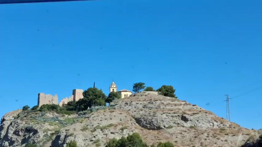 The video captures a view from a vehicle, showing a hilltop with ancient ruins and a church under a clear blue sky. The ruins, appearing to be a castle, are located on the left side of the hilltop, with stone walls and structures visible. To the right of the ruins is a yellow church with a tall steeple. The hilltop is covered with sparse vegetation, including green bushes and agave plants. The terrain is rocky and arid. The vehicle appears to be moving, as evidenced by the slight camera shake and the changing perspective of the scene. There are no people visible in the video. The overall impression is of a historic site in a rural, sun-drenched environment.