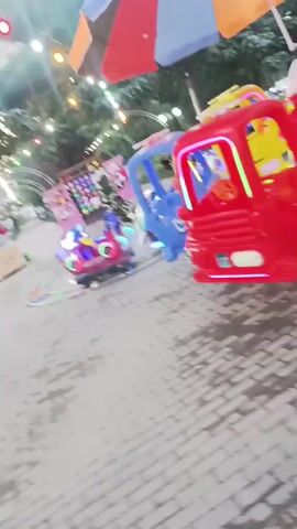 The video shows a park with amusement rides. In the foreground, there are several colorful, small amusement park vehicles, including red, blue, and yellow cars. Some of these vehicles are spinning and have flashing lights. In the background, there are more amusement rides, including a carousel with smaller vehicles. There are also people walking around, and string lights are visible in the trees. The ground is paved with bricks. The overall atmosphere appears to be festive and lively.