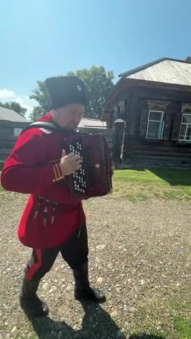 A man in a red traditional outfit and a black fur hat plays an accordion. He is standing outside on a gravel surface, with a wooden house and trees in the background. The man is looking at the camera and appears to be smiling. The sky is blue and clear, indicating a sunny day.