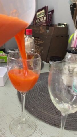 Here's a brief description of the video:
A bright orange liquid is poured from a clear container into a wine glass on a white table with a gray, circular placemat. Other items visible in the background include a bag of tortilla chips, a bottle with a red cap, a jar labeled "Coconut Butter Creamy Pure," a box, and a bouquet of dried flowers with ribbons. The camera angle shifts slightly during the pouring.