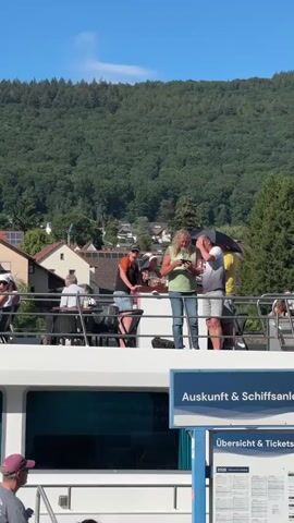 The video shows a group of people on the deck of a boat. The boat appears to be docked at a pier, as indicated by the "Auskunft & Schiffsanleger" sign, which translates to "Information & Ship Dock". In the background, there is a hillside covered in green trees and houses. The sky is clear and blue.

On the boat deck, several individuals are visible. In the foreground, a person with a pink hat and a gray t-shirt is walking down stairs. To their right, a person with dark hair and sunglasses is standing. Further up on the deck, a man with a long, blonde ponytail and a green shirt is holding a phone and looking at it. He is standing next to another man, partially obscured by an umbrella, who is also looking down. To the left of the blonde-haired man, a person wearing a black t-shirt and blue shorts is leaning over a railing, talking to someone. Another group of people are seated at tables on the left side of the deck. The overall atmosphere appears to be relaxed and social, likely a tourist excursion.