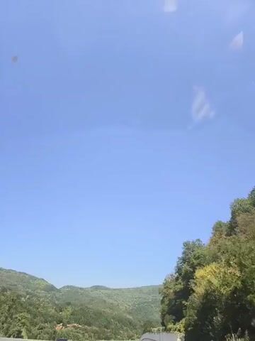 A car is driving on a highway. The sky is blue and clear. There are trees and mountains on the side of the road. The car is black and has its headlights on. The road is made of asphalt and has white lines marking the lanes. There is a concrete barrier on the right side of the road. The trees are green and lush. The mountains are covered in vegetation.