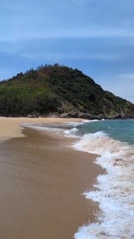 The video shows a serene beach scene. The foreground is dominated by a sandy beach with gentle waves lapping at the shore. The water is a clear blue-green, and the waves create a white foamy border as they recede. In the background, a lush green, tree-covered hill rises prominently, its slopes meeting the sky. The sky itself is a pale blue, with a few wispy clouds scattered across it. The overall impression is one of natural beauty and tranquility.