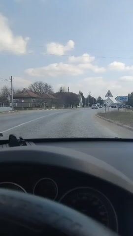 The video shows a view from the driver's seat of a car moving along a road in what appears to be a small town or village. The road is asphalt and has a few other vehicles in the distance, including cars and trucks. Buildings line the sides of the road, mostly single-story structures with traditional roofs. Trees are also present, with bare branches suggesting it might be late fall or winter. There's a billboard on the right side of the road with the words "BETWILD COMING SOON" written on it. The sky is light blue with some scattered clouds. The steering wheel of the car is visible in the foreground. The dashboard is also visible.