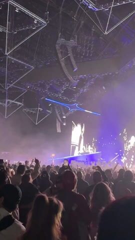 A music event unfolds inside a large, high-ceilinged structure with a visible geometric framework overhead. A DJ is perched on a raised platform, visible amidst a throng of concertgoers. The crowd, packed tightly together, raises their hands and films the event on smartphones. Lighting displays are prominent, including blue laser-like beams and spotlights piercing through the haze. Hanging geometric shapes are suspended from the ceiling, enhancing the visual spectacle. The atmosphere is energetic, with music booming through the venue.