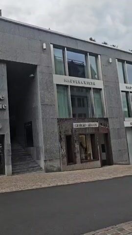 The video shows a street scene with a building featuring a prominent entrance labeled "Palace Galerie" with the letters "OT1" beneath it. To the right, the building has windows and displays for various luxury brands, including "HAUSER&WIRTH," "GIORGIO ARMANI," and "LOUIS VUITTON." People are seen walking along the sidewalk, and a white SUV is driving on the street. The architecture is modern, with gray stone facades and large glass windows. The sky is overcast.
