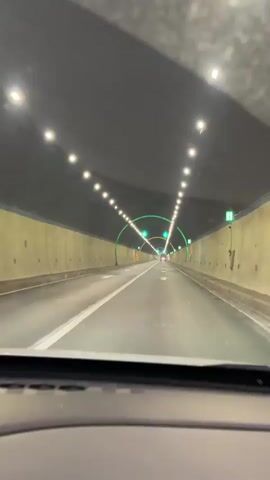 A car is driving through a brightly lit tunnel. The tunnel has rows of overhead lights that converge in the distance. Green arches run over the lanes, adorned with arrows pointing forward. The road has painted lane markers and directional arrows. The tunnel walls are a light color. The view is from inside the car, showing the dashboard.