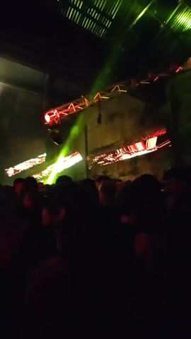 The video shows a dark, crowded space that appears to be a music venue or concert. The crowd is mostly silhouetted, but some individuals are visible. Bright green spotlights move around the stage, and there are other lights that appear to be orange or red. The environment looks like an industrial space, with visible beams and ceiling structures. The audience seems to be enjoying the performance, with some raising their hands.