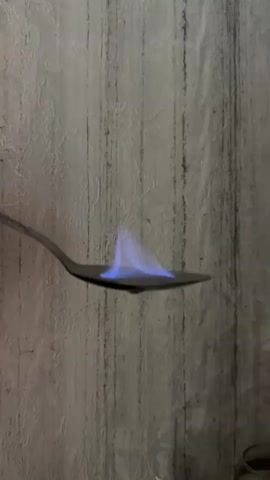 A hand holds a metal spoon over a textured, light-colored wall. A blue flame flickers on the surface of the spoon. In the background, out of focus, are several clear glass objects, possibly drinking glasses, and a small white object rests on a surface below the spoon.