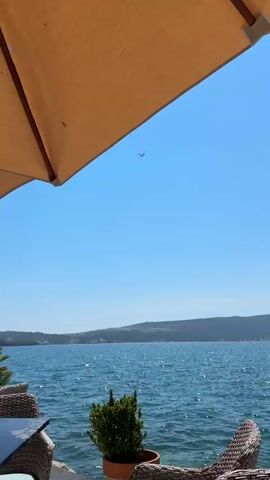 The video captures a serene waterside scene under a bright, clear sky, suggesting a sunny day, likely late morning or early afternoon. The foreground is partially obscured by the edge of a large, light brown umbrella, casting a soft shadow. Below the umbrella, wicker chairs and a table are visible, hinting at an outdoor seating area, perhaps a cafe or restaurant. A potted green plant adds a touch of nature to the setup.

The main focus beyond the immediate seating area is a wide expanse of shimmering blue water, reflecting the sunlight. Across the water, a distant, hilly coastline is visible, dotted with buildings that suggest a town or settlement. The sea appears calm with gentle ripples. High in the sky, a small airplane is flying, its silhouette clearly defined against the vast blue. Further out on the water, a small boat can be seen in the distance, adding a subtle sign of activity.

The overall mood is peaceful and relaxed, evoking a sense of vacation or leisure. The clear weather and the presence of the sea and coastline strongly suggest a coastal location. The mention of Tivat, Montenegro, confirms this is a seaside resort or town. There is no visible text in the media. The activity taking place is the observation of this picturesque view, with the subtle movement of the airplane and the boat adding a sense of life to the otherwise tranquil scene.