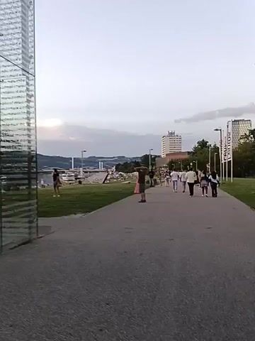 The video shows a paved walkway in a park with grass on the left. A person stands on the grass to the far left, wearing a dark top and shorts, facing away from the camera. In the middle ground, a path leads towards a group of people walking away from the camera. A man in a green t-shirt and shorts stands on the path, with his hands on his head. Further down the path, a group of people, mostly wearing white clothing, walk away from the camera. In the background, there is a body of water, a bridge, buildings, and hills under a cloudy sky. The lighting suggests it is late afternoon or early evening.